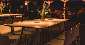 Das Bild zeigt einen Teil der Eventlocation LAKESIDE während eines Winter-Events. Draußen ist es dunkel, die Location ist daher in angenehmen Goldtönen erleuchtet. Im Vordergrund steht ein weißer Stehtisch mit sechs weißen Barstühlen.