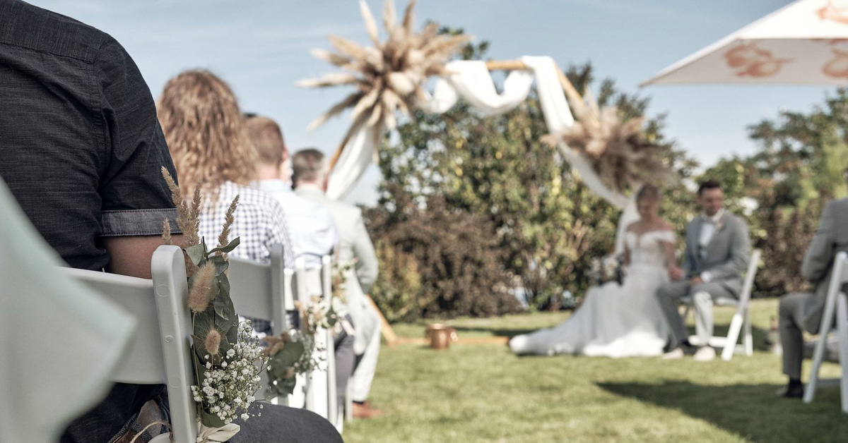 Blick auf den Traubogen im Garten des LAKESIDE aus Sicht eines der Gäste. Das Hochzeitspaar sitzt seitlich des Bogens.