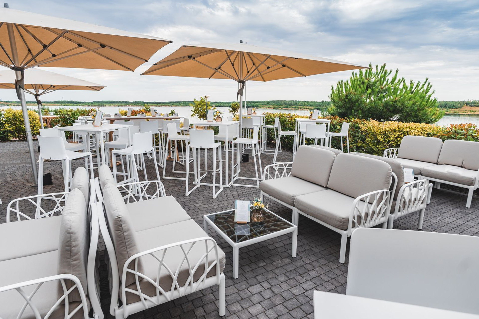 Die vordere See-Terrasse der LAKESIDE Eventlocation bei Leipzig: bequeme weiße Sofas mit grauen Polstern, im Hintergrund moderne weiße Stehtische und Barhocker, Sonnenschirme sowie Bepflanzung und ein Teil des Sees.