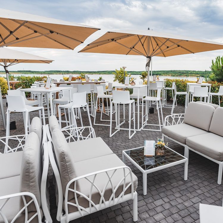 Seeterrasse der LAKESIDE Eventlocation mit weißen Loungemöbeln, Sonnenschirmen und dem See im Hintergrund