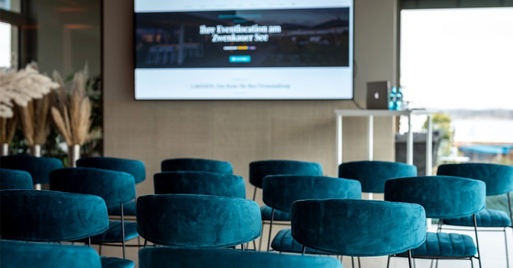 Setup für Firmenveranstaltungen im LAKESIDE: pretrolfarbene Stühle in Reihe vor großem Display an der Wand