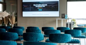 Setup für Firmenveranstaltungen im LAKESIDE: pretrolfarbene Stühle in Reihe vor großem Display an der Wand