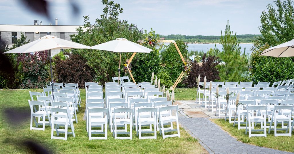 Blick über den Garten des LAKESIDE mit Trauungs-Setup aus Traubogen und weißen Stühlen in Richtung See.