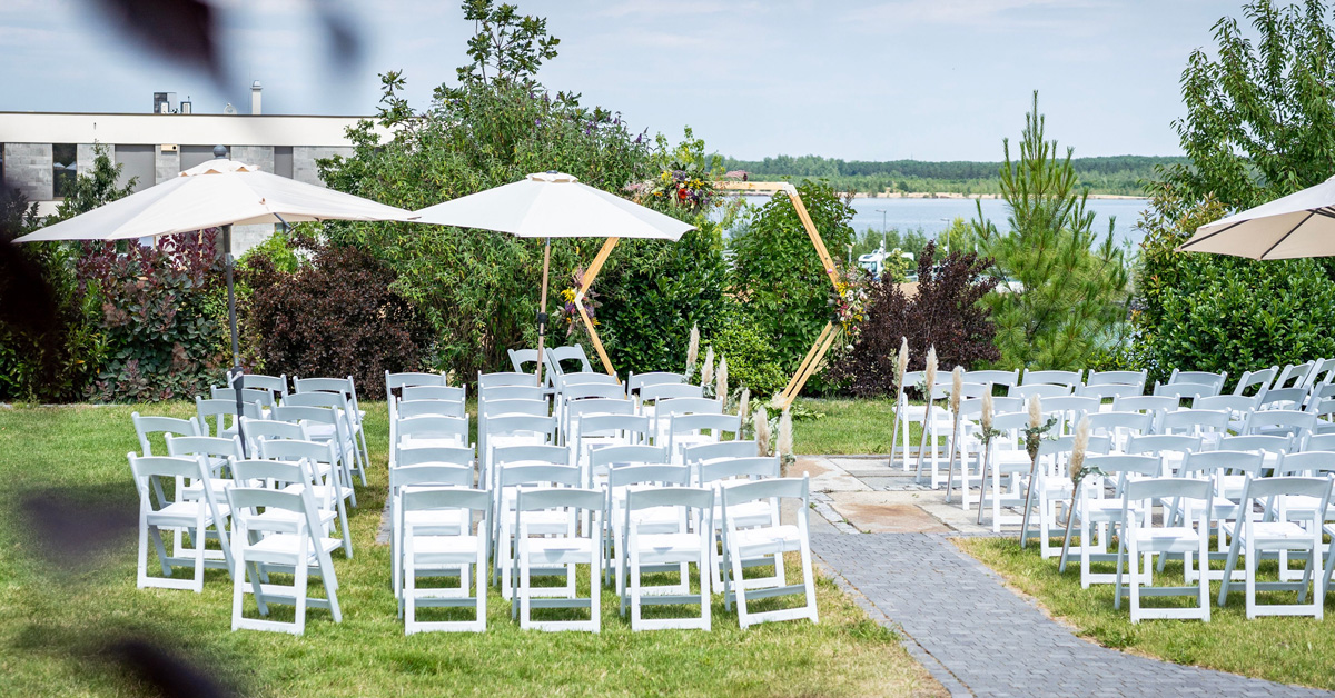 Blick über den Garten des LAKESIDE mit Trauungs-Setup aus Traubogen und weißen Stühlen in Richtung See.