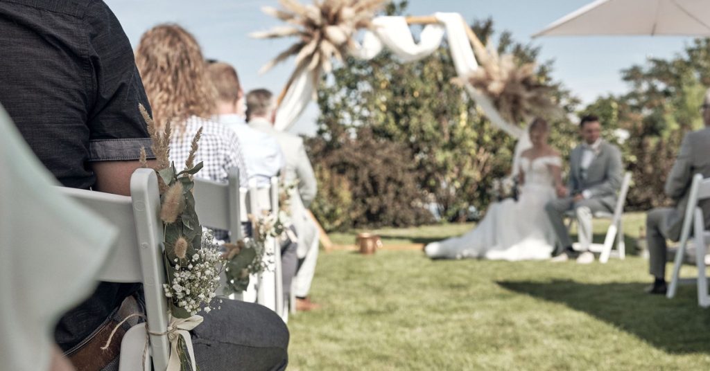 Trauung-Setup in der Hochzeitslocation LAKESIDE: Gäste sitzen auf geschmückten weißen STühlen, im Hintergrund sitzt das Brautpaar seitlich vor dem geschmückten Traubogen