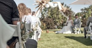 Trauung-Setup in der Hochzeitslocation LAKESIDE: Gäste sitzen auf geschmückten weißen STühlen, im Hintergrund sitzt das Brautpaar seitlich vor dem geschmückten Traubogen
